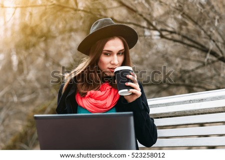 Stylish beautiful girl in hat sitting on bench in sunny autumn park. Drink warm coffee and working outside with notebook in open air