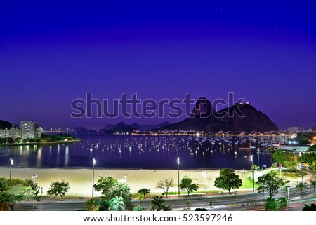 Night photography of Sugar Loaf mountain and Botafogo Bay in Rio de Janeiro, Brazil. This picture shows the beach, trees, boats, streets and city lights.