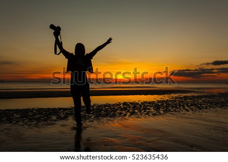 Silhouette woman photographer walking on beach with hand holding camera at sunrise.