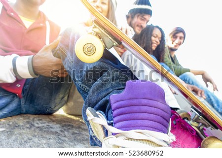 Group of happy friends having fun outdoor in urban city park with sun back light - Young youth people socializing - Leisure and friendship concept - Focus on skateboard wheel - Warm filter