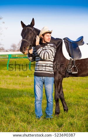 Beautiful strong man cowboy nearby black horse. Has happy  face, striped pullover, blue jeans, hat. Has sport body. Portrait nature. People and animals. Equestrian. Amazing landscape.