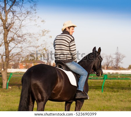 Beautiful strong man cowboy riding black horse. Has happy  face, striped pullover, blue jeans, hat. Has sport body. Portrait nature. People and animals. Equestrian. Amazing landscape.