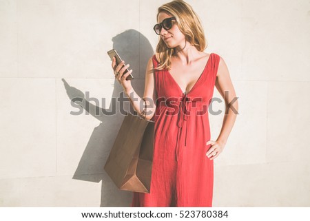 Summer sunny day,young woman in sunglasses and red dress standing outside and looking on screen of smartphone,in her hand she holds shopping bag.In background white wall.Girl using gadget.Film effect.