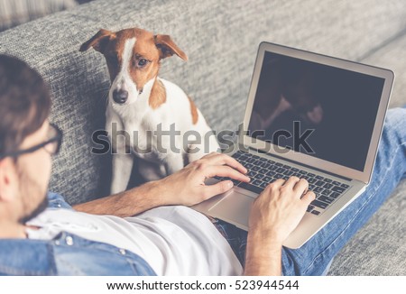 Handsome man in eyeglasses is using a laptop while lying on couch at home. Cute dog is looking at his guardian Royalty-Free Stock Photo #523944544