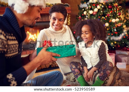 
Smiling lovely girl receiving Christmas present from parents, happy family near fireplace