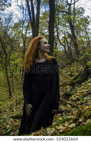 Young beautiful red-haired girl dressed like a witch in a black clothes, with long hair and owl tattoo, looks like a fox, smiling and sitting on a large log with moss. Forest background. Green.