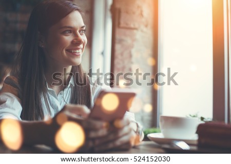 Smiling woman in cafe using mobile phone and texting in social networks, sitting alone Royalty-Free Stock Photo #524152030
