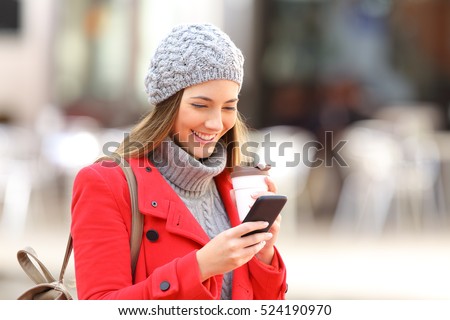 Happy fashion woman using a smart phone and holding a take away coffee in the street in winter
