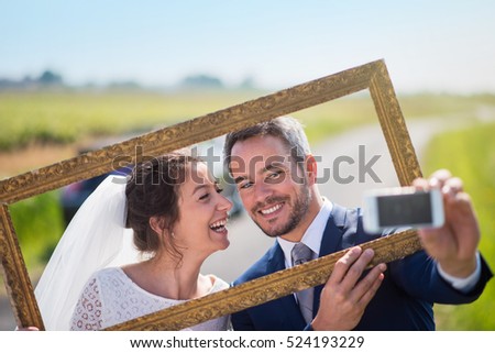 young married couples having fun to take pictures of themselves through an empty frame. this is the husband who holds his mobile and takes a selfie.they are on a country road