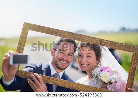 young married couples having fun to take pictures of themselves through an empty frame. this is the husband who holds his mobile and takes a selfie.they are on a country road