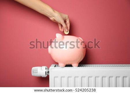 Savings concept. Female hand putting coin into piggy bank which standing on heating radiator with temperature regulator on pink background