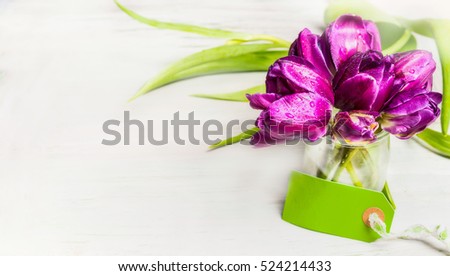 Tulips bunch in glass jar with water and empty tag on light background, banner. Spring and greeting flowers concept.