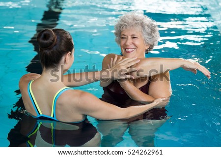 Young trainer helping senior woman in aqua aerobics. Senior retired woman staying fit by aqua aerobics in swimming pool. Happy old woman stretching in swimming pool with young trainer. Royalty-Free Stock Photo #524262961