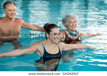 Smiling fitness class doing aqua aerobics in swimming pool. Smiling young woman with senior couple stretching arms in swimming pool. Fit mature man and old woman exercising in swimming pool. Royalty-Free Stock Photo #524262967