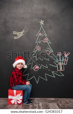 Picture of happy child wearing hat sitting on big gift near Christmas tree drawing on blackboard. Looking at camera.