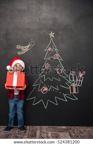 Picture of joyful screaming child wearing hat holding a big gift near Christmas tree drawing on blackboard. Looking at camera.