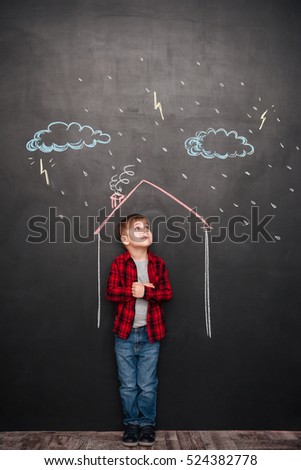 Picture of cute little kid standing in house on the chalkboard with drawings of a rain. Looking up to drawings.