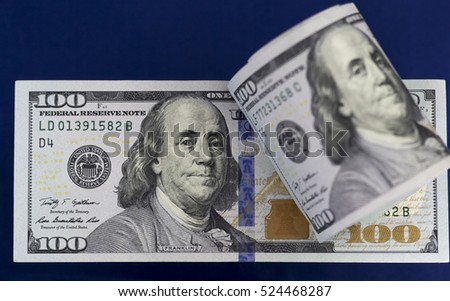 Rolled hundred us dollars banknote is lying on another flat hundred american dollar bill. On dark blue background.
