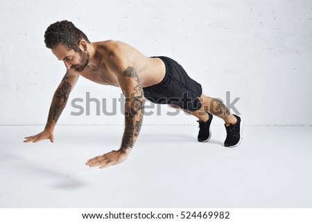 Strong brutal tattoed and ripped male athlete shows how to do explosive in out push-ups, isolated on white spacious room, mid position, pushing in air