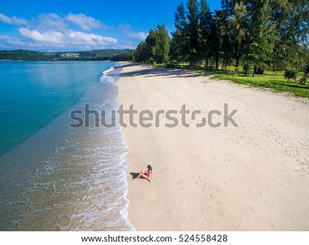 Young woman in a pink bikini sitting on the sand near turquoise sea. Top view. Bangtao beach, Phuket, Thailand. Aerial Shooting.

