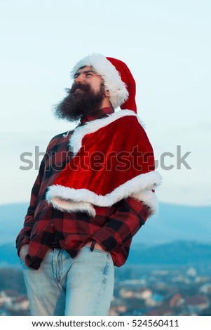 young handsome smiling bearded bad santa claus man with long beard in checkered shirt jeans and red new year hat in christmas or xmas coat outdoor on blue sky background