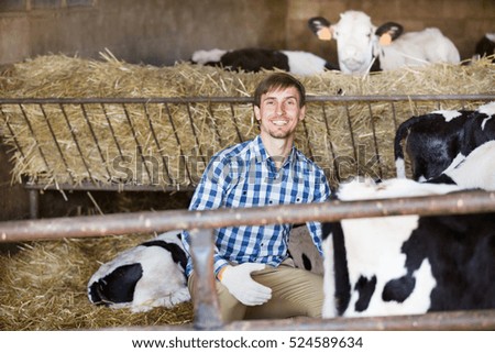young male farmer sitting with cows in farm hangar and smiling