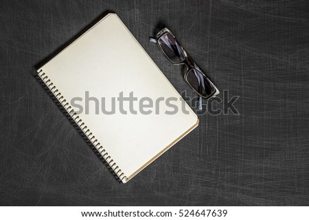 Single simple empty white notebook with a blank for drawing or writing and eyeglasses are on a Desktop from black chalkboard. Top view. Mockup. Flat lay