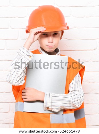 young cute builder boy in orange uniform in hard hat or helmet holding paper on white brick wall background