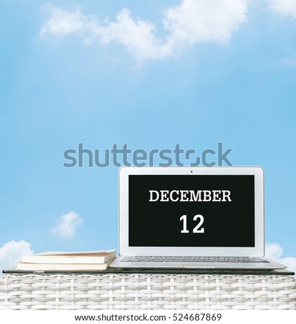 Closeup computer laptop with december 12 word on the center of screen in calendar concept on blurred wood weave table and book on blue sky with cloud textured background with copy space