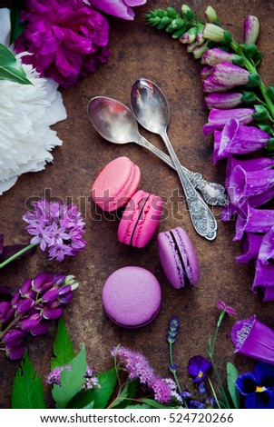 Purple and pink French macaroon cookies, a spoon and a variety of flowers on a wooden background