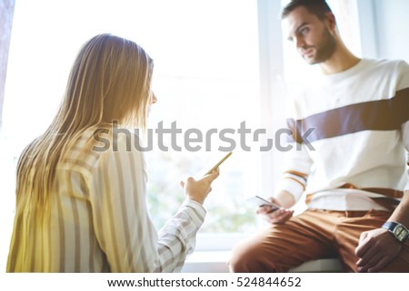 Male and female office workers watching popular video in network and sharing photo files via cellulars. Two coworkers recommending the convenient to each other using smartphone programs