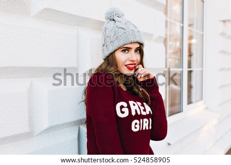 Portrait pretty girl with long hair in marsala sweater on grey background outside. She wears knitted hat, keeps finger on red lips and smiling to camera