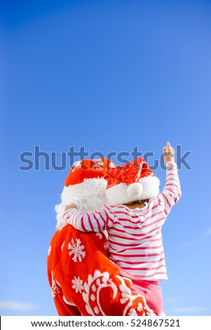 Happy Merry Christmas. Child hugging Santa Claus father together wearing red hats over sunny blue sky background outdoors. Back view