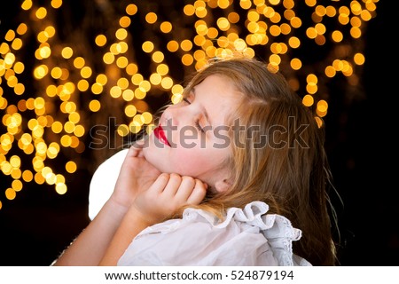 A blissful, young girl with blond hair sits in front of a starry, Christmas light background with her chin in her hands and head tilted back.  She has closed eyes and a closed mouth smile.