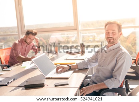Closeup picture of handsome businessman sitting in front of laptop computer and looking at camera while his colleague working on background in board room in office.