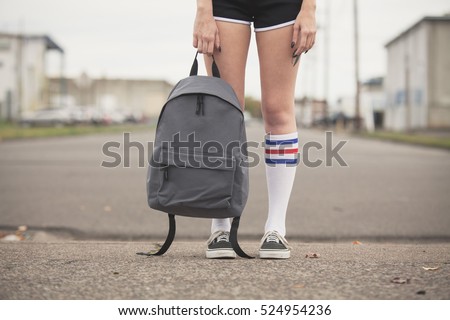 Girl holding generic backpack wearing black shorts and tube sock with tennis shoes standing on vacant street. 