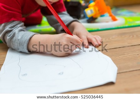 Little child drawing with pencil on white paper. Close up of toddler playing on floor. 