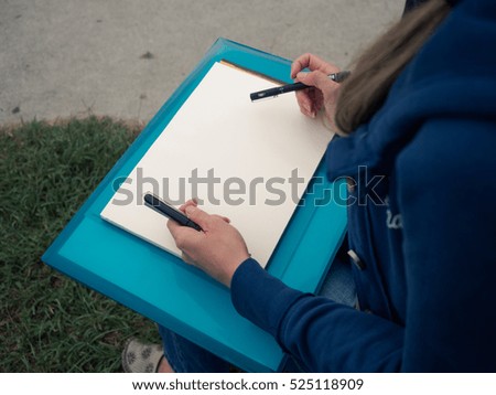 Picture of the girl draws on the clean sheet of paper. Female has blue suit on draws on the paper with felt pen. Clean sheet of paper on the girl's knees. The girl draws on the paper.