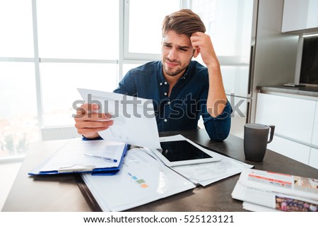 Young confused man analyzing finances at home while holding head with hand and looking at documents. Sitting near table with tablet. Royalty-Free Stock Photo #525123121