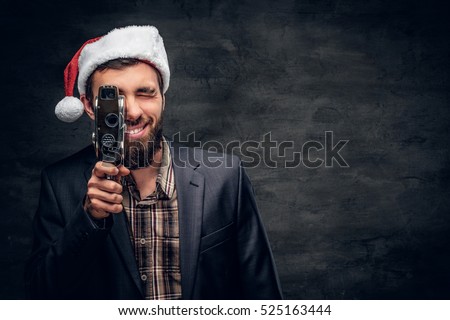Bearded male in Santa's hat holds the old 8mm video camera on grey vignette background.