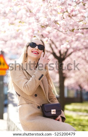 Smiling lady in beig coat sits under pink blooming tree