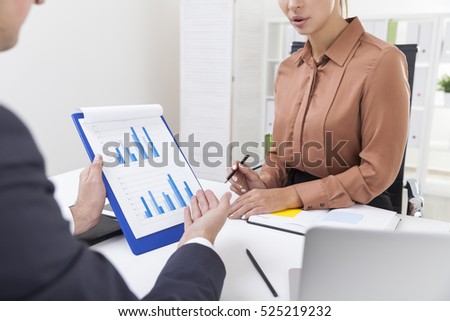 Man and a woman in office. They sit at a table and work with a blue diagram attached to a notepad. Overt the shoulder shot.