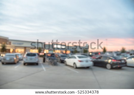 Blurred image exterior view of modern shopping center in Humble, Texas, US at sunset. Mall complex with row of cars in outdoor uncovered parking lots and bokeh light of retail store in background.