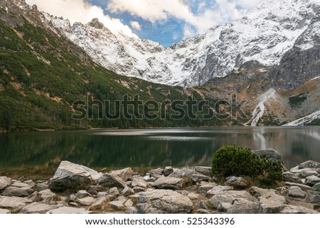 The landscape of  lake surrounded by snow mountains with  green fir-trees  and  blue sky with white clouds and big stones in the foreground