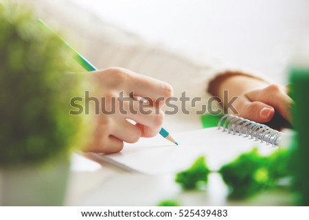 Close up of girl's hand writing in spiral notepad on desktop with decorative plant