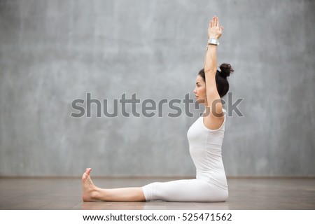 Side view portrait of beautiful young woman working out against grey wall, doing yoga or pilates exercise without mat on wooden floor. Model sitting in Dandasana, Staff pose. Full length Royalty-Free Stock Photo #525471562