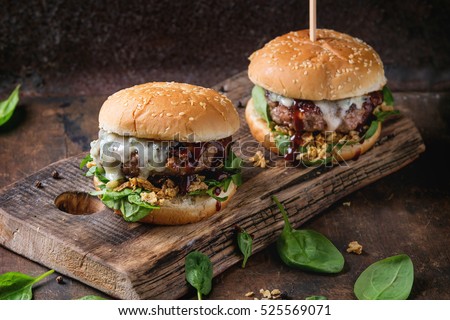 Two hamburgers with beef burger cutlet, fried onion, spinach, ketchup sauce and blue cheese in traditional buns, served on wood chopping board over dark wooden background.