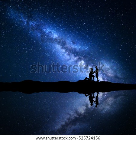 Milky Way. Silhouettes of a man making marriage proposal to his girlfriend on the mountain near the lake with starry sky reflection in water. Night landscape. Couple. Blue milky way with people. Space