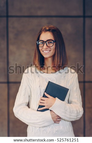 Young beautiful smiling girl in stylish glasses and a warm white sweater with sketchbook on the background of a concrete wall