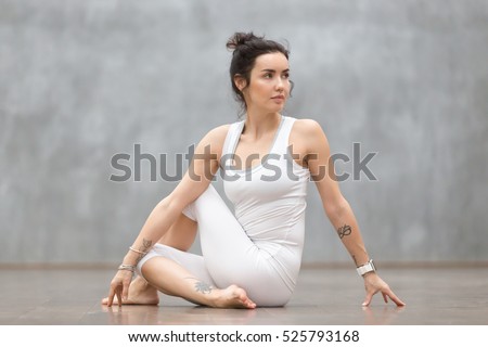 Beautiful young woman with floral tattoo working out against grey wall, doing yoga or pilates exercise. Girl sitting in Half lord of the fishes, Ardha Matsyendrasana pose. Full length Royalty-Free Stock Photo #525793168